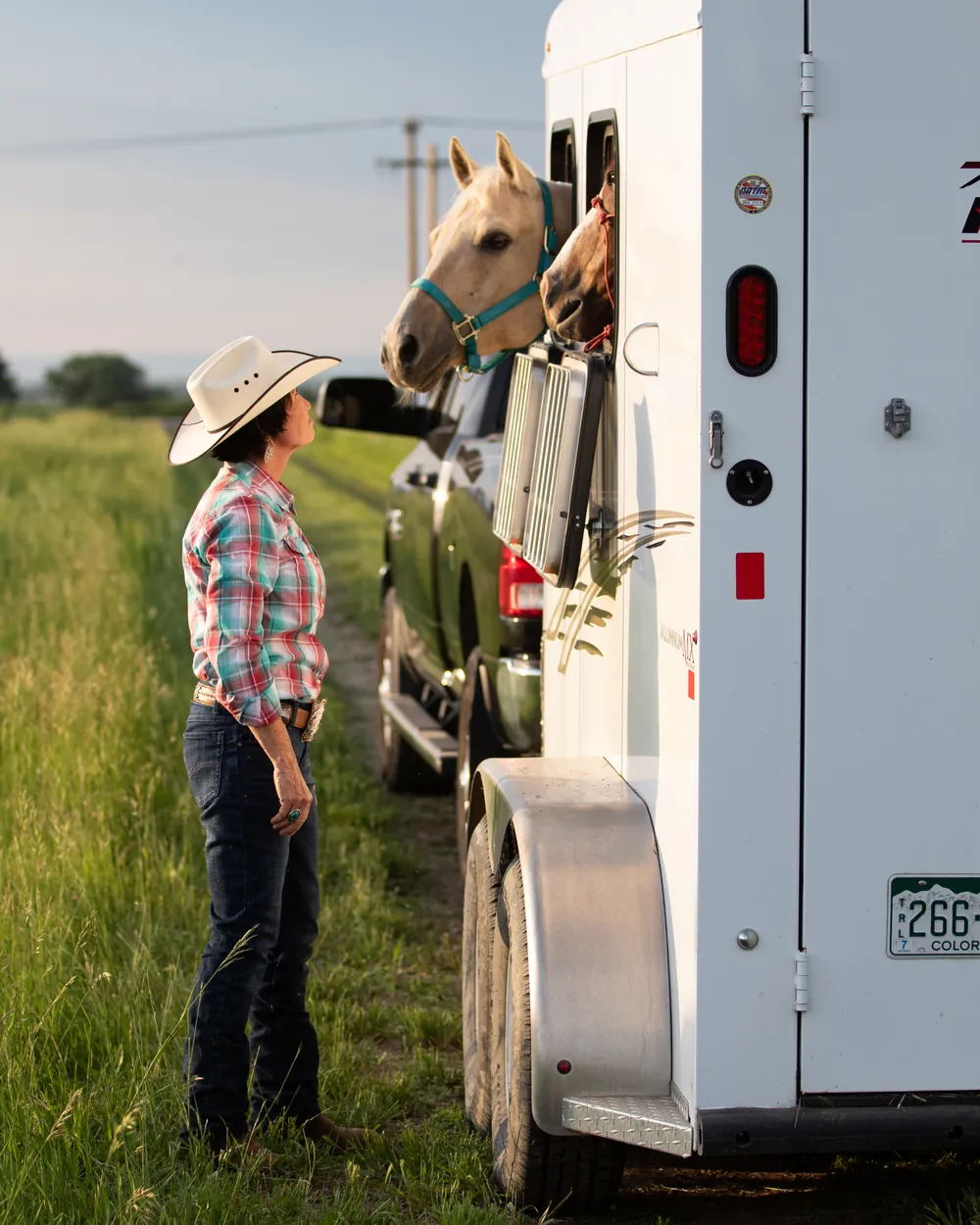 Rider in a cowboy hat greeting two horses in a trailer at golden hour